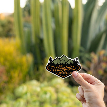 In front of tall cactus and golden light, someone holds The Mountains Are Calling sticker by one corner.