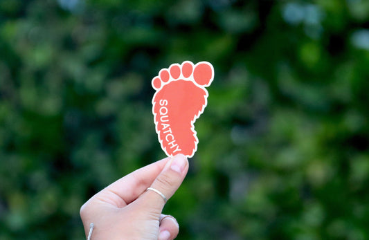 Pulled back wider, the Squatchy Logo Sticker sits small between two fingertips with a hedge filling the frame.