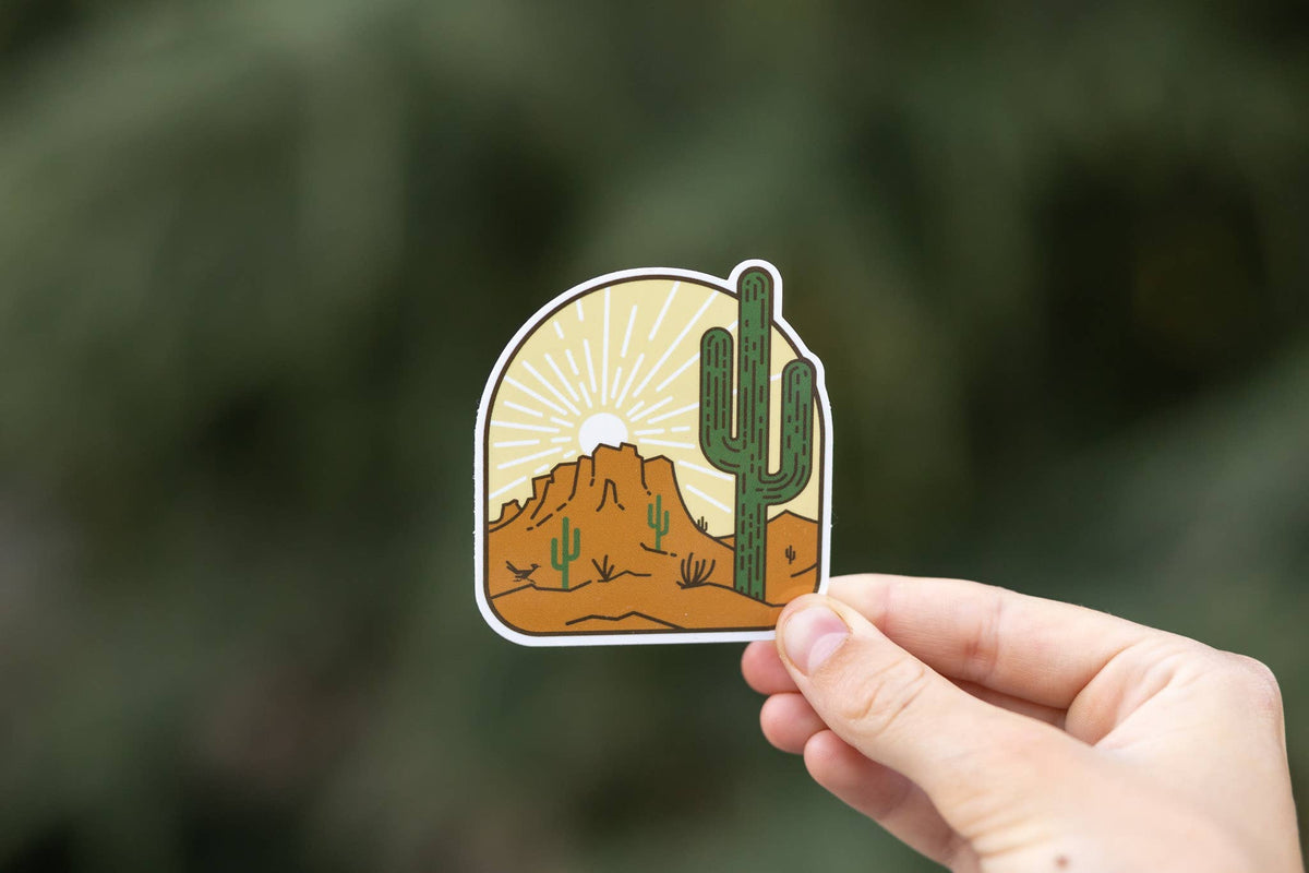 A hand holds the Desert Landscape sticker against blurred green foliage, saguaro and butte under a rising sun.