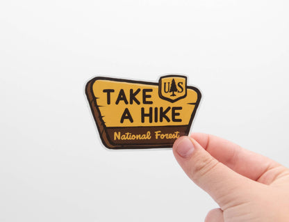 A hand holds the yellow Take a Hike sticker, shaped like a National Forest trail sign, against a white wall.