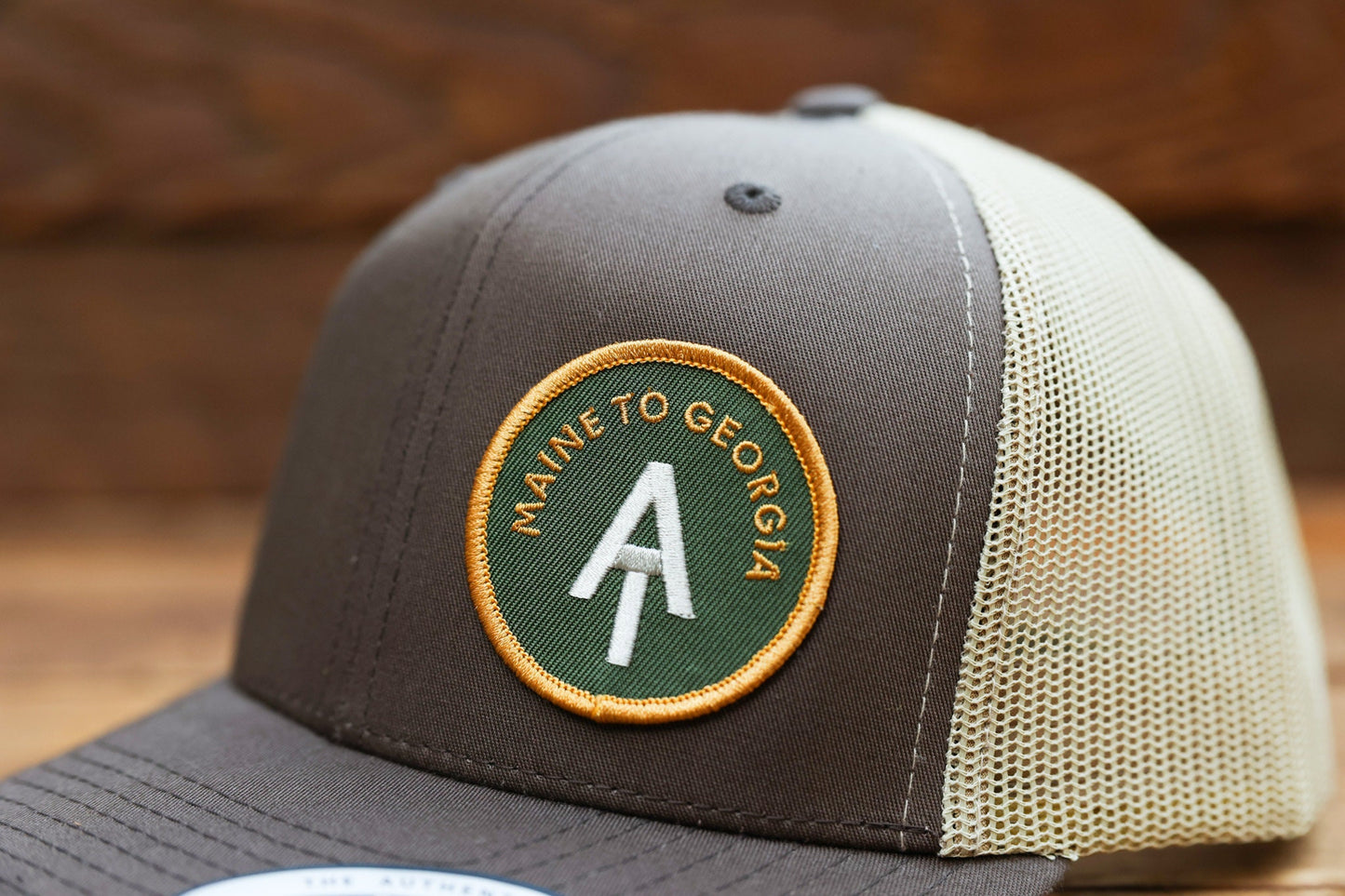 Tight crop of the brown Appalachian Trail trucker hat, patch letters curving around a white trail marker.