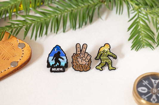 Three pins in a row, Believe, the furry peace hand and the Sasquatch, with cedar and a compass nearby.