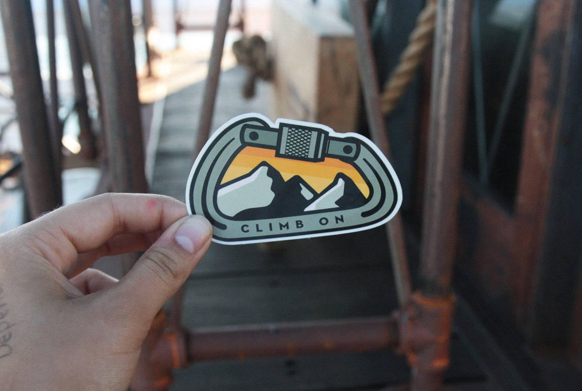 On a pier with rusted pipes behind, a hand raises the Climb On Carabiner sticker in low sun.