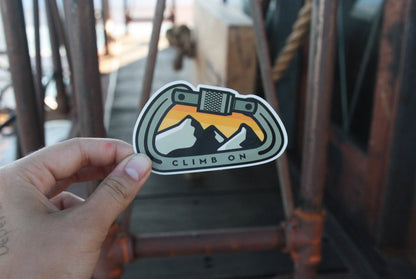On a pier with rusted pipes behind, a hand raises the Climb On Carabiner sticker in low sun.