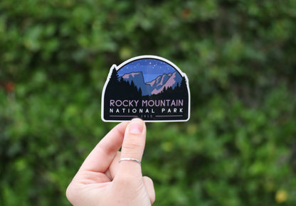 Held further back from the same green shrubs, the Rocky Mountain National Park sticker sits small in the frame.