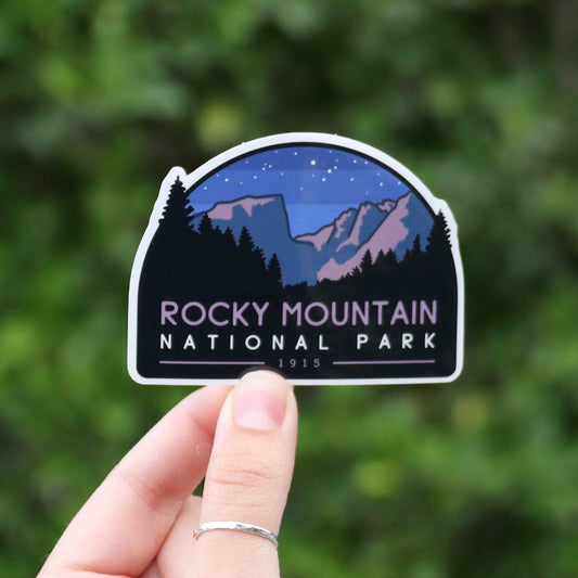 Held further back from the same green shrubs, the Rocky Mountain National Park sticker sits small in the frame.
