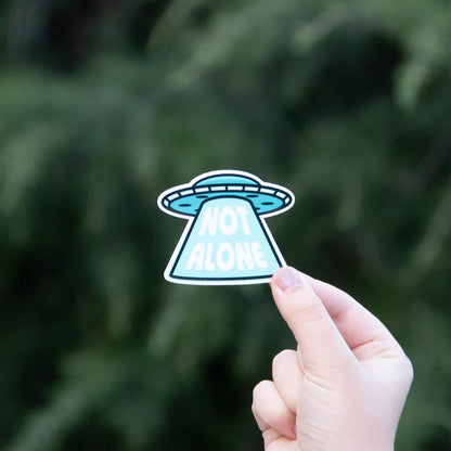 A hand holds up the Not Alone UFO sticker, its blue saucer and beam of lettering bright against dark pine.