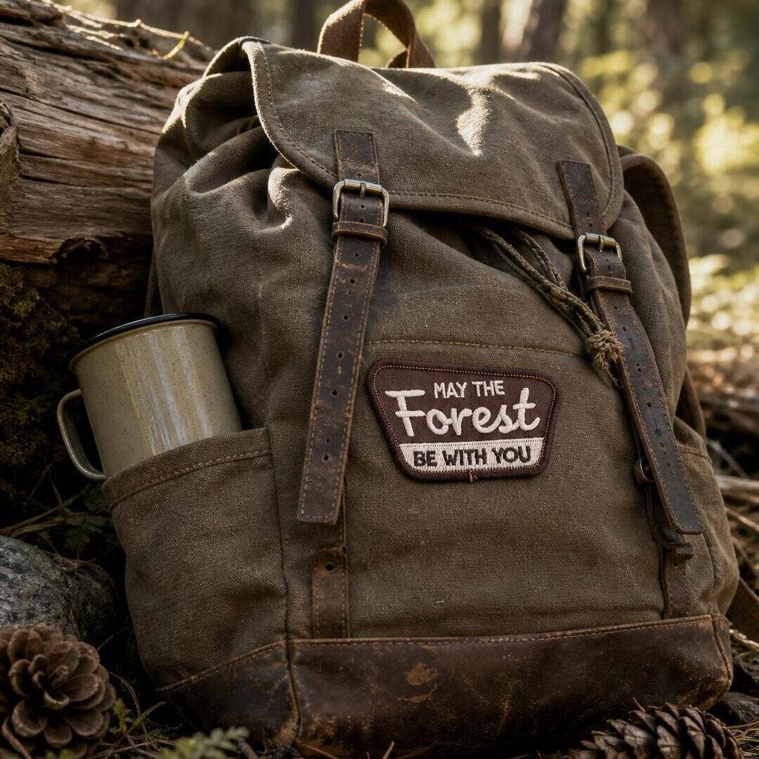 Stitched to a waxed canvas backpack beside an enamel mug, the May The Forest Be With You patch sits in the woods.