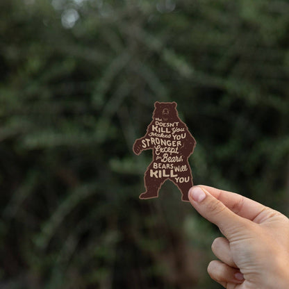 Fingertips pinch the lower edge of the Bears Will Kill You sticker in front of dark conifer branches.
