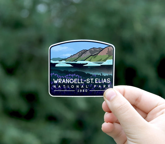 A hand holds up the Wrangell St. Elias National Park sticker, its glacier and peaks bright against blurred greenery.