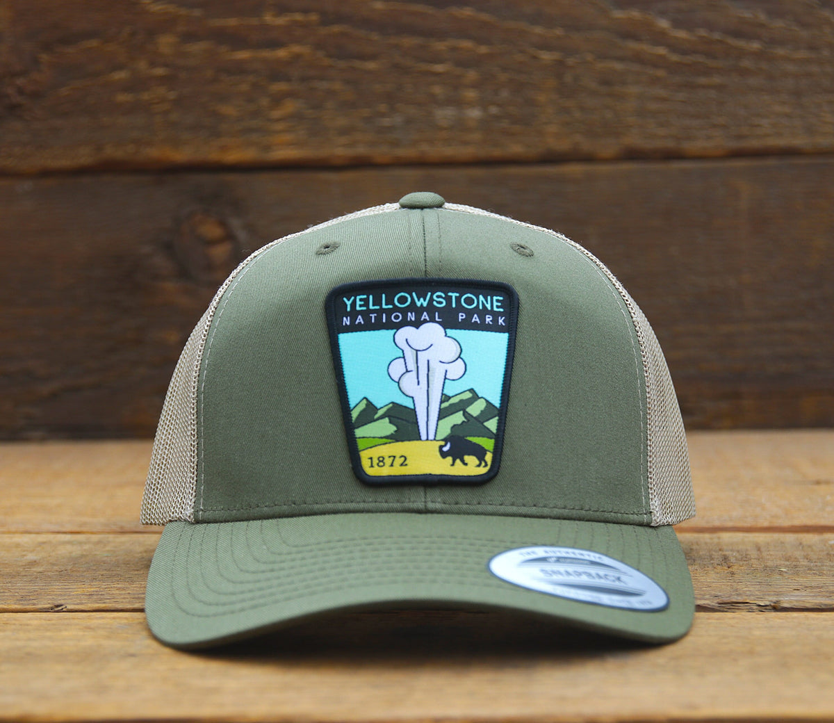Olive Yellowstone National Park trucker hat squared up on weathered boards, geyser and bison patch centered.