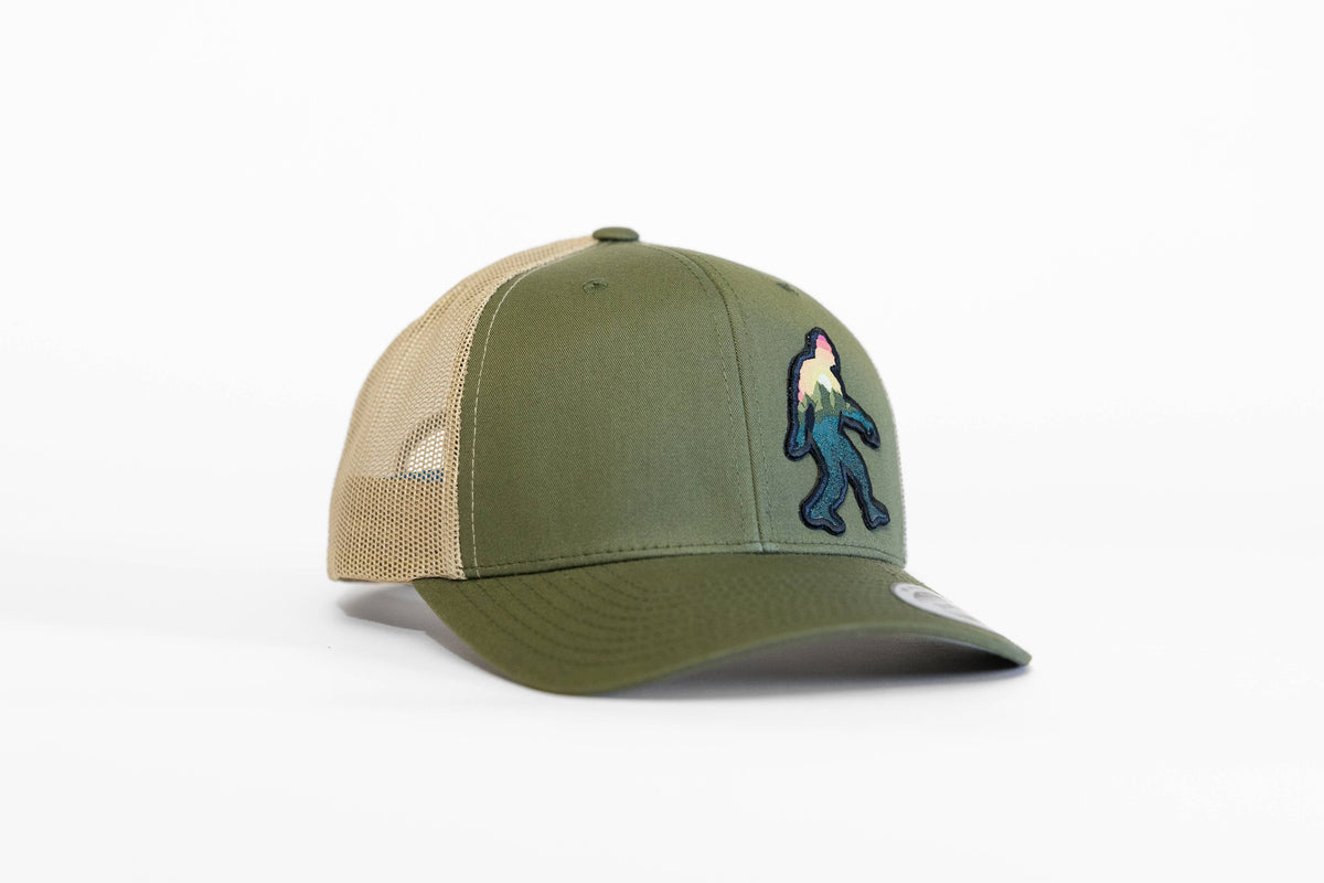 Olive green Bigfoot Adventure trucker hat with tan mesh sides and the mountain filled Sasquatch patch on the crown.