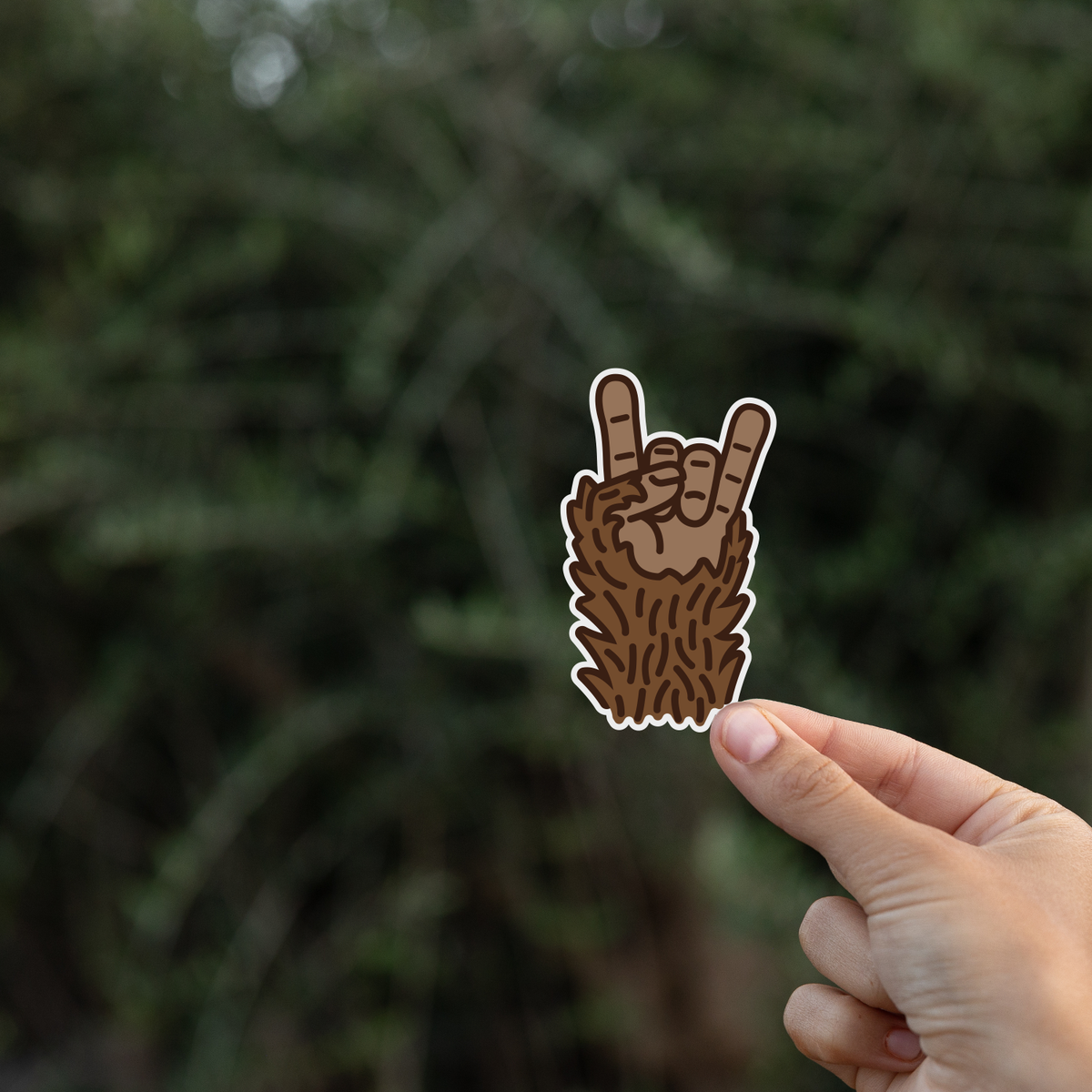 Pinched at one corner, the Bigfoot Rocks sticker stands against dark evergreen branches.