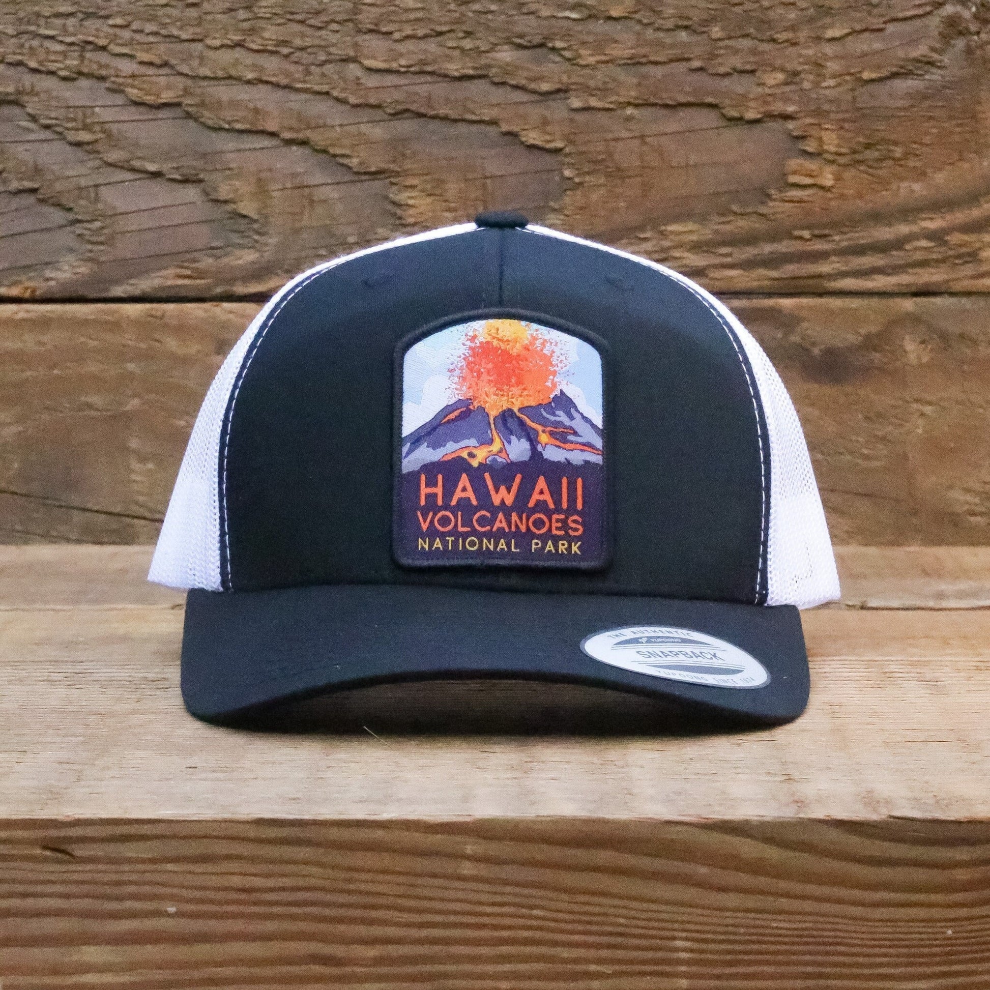 Centered against weathered barn wood, the black and white Hawaii Volcanoes trucker hat faces forward.