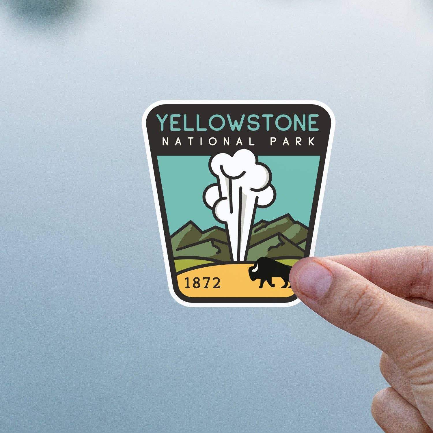 Against a soft blue and grey background, a thumb and finger hold the Yellowstone National Park sticker.