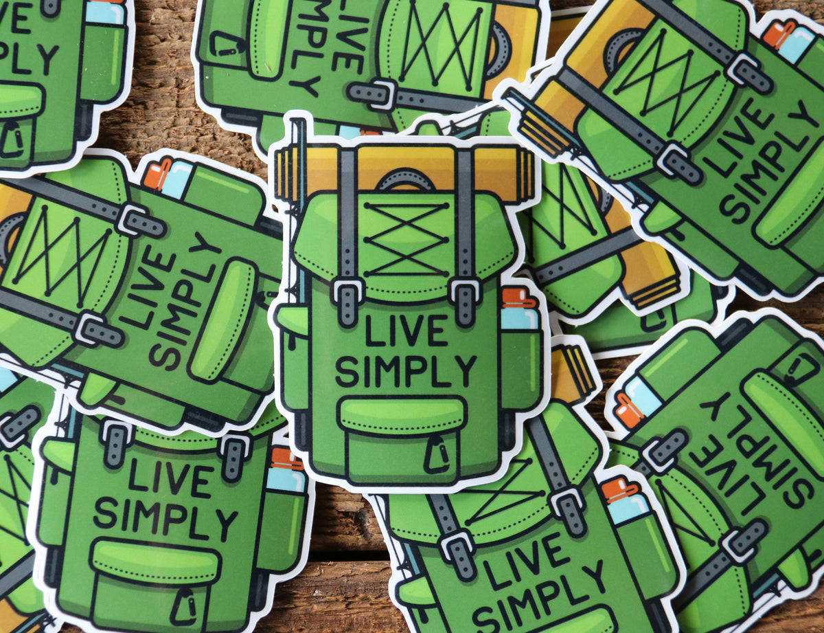 Scattered across weathered planks, a pile of Live Simply backpack stickers overlaps with one centered on top.
