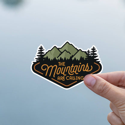Two fingers hold The Mountains Are Calling sticker against a soft grey and blue background.