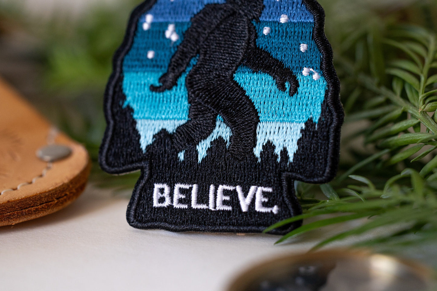 Up close among greenery and leather, the white BELIEVE letters on the Bigfoot patch read sharp and raised.