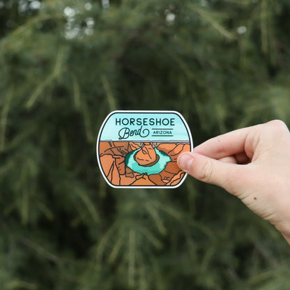 A hand holds the Horseshoe Bend sticker against blurred evergreen branches, showing the red canyon and green river.