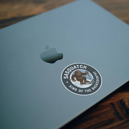A pale blue laptop lid on dark wood carries the Sasquatch King of the Backcountry sticker below the Apple logo.