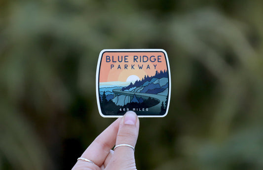 Pinched between two ringed fingers, the Blue Ridge Parkway sticker sits small in a wide frame of soft greenery.