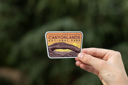 Against a darker blurred evergreen, fingers hold the Canyonlands National Park sticker with its yellow sun under the arch.