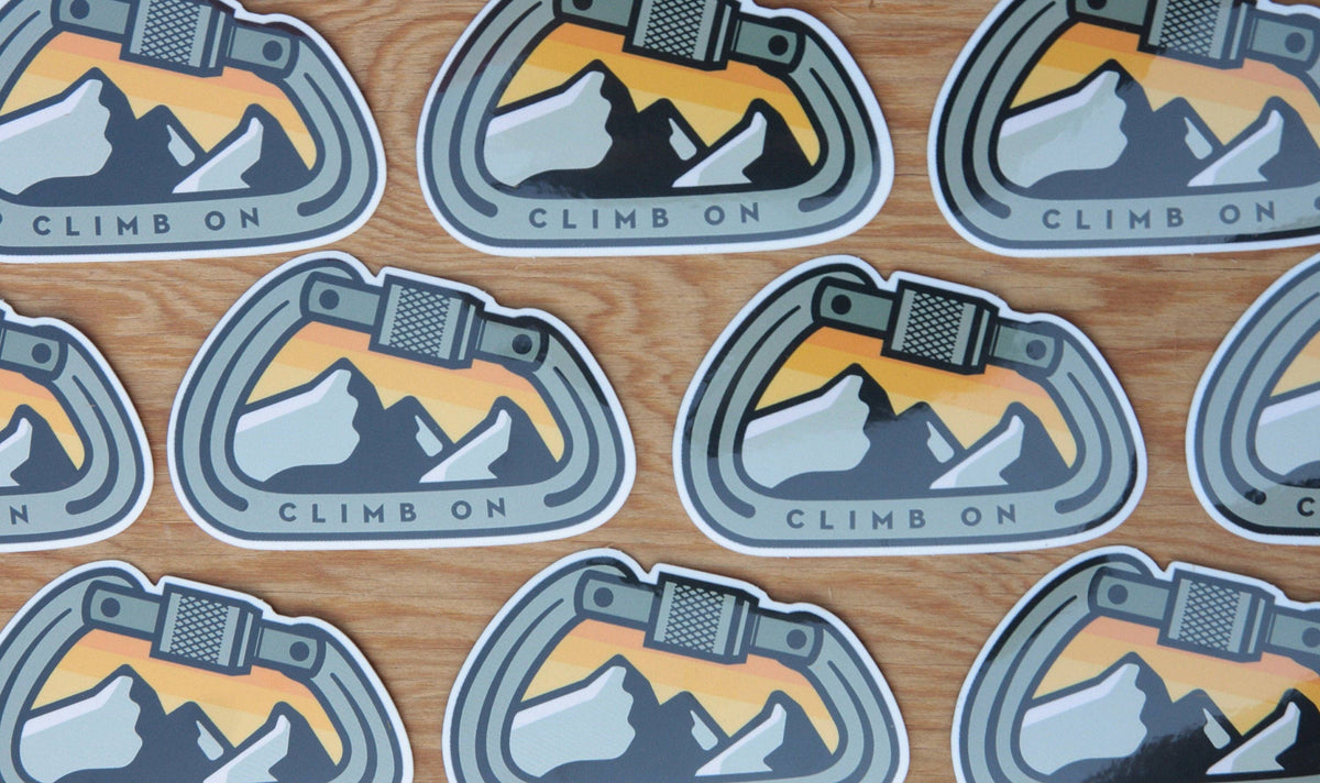 Rows of Climb On Carabiner stickers lie spread across a plywood surface in a loose grid.