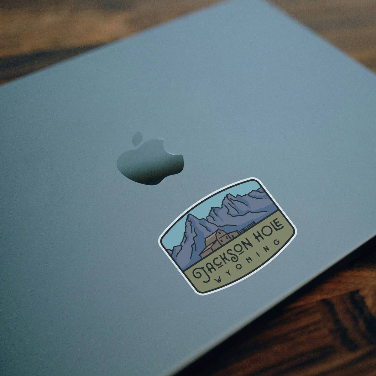 Angled across a silver laptop lid near the apple logo, the Jackson Hole Wyoming sticker catches soft light.