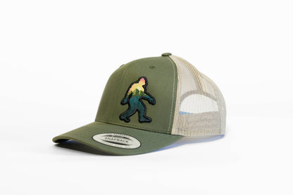 The olive and khaki Bigfoot Adventure trucker hat sits at an angle with its snapback sticker still on the brim.