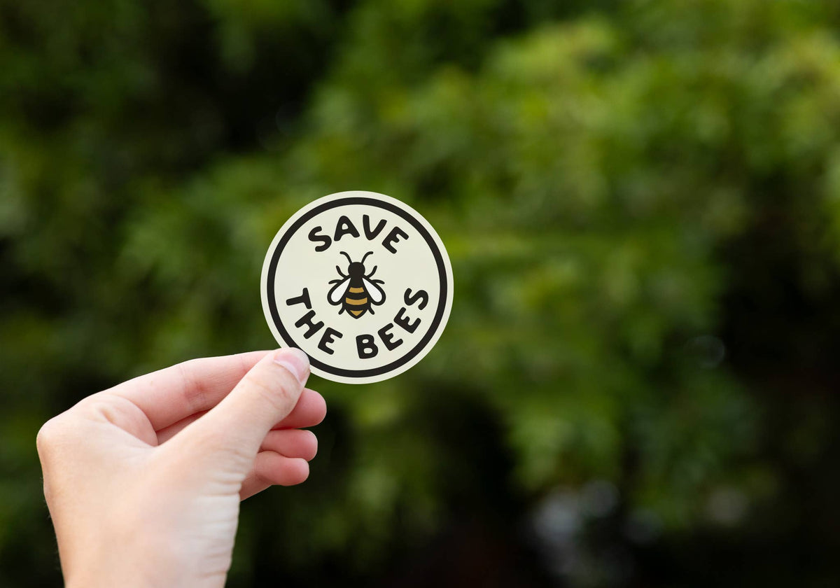 A hand holds the round Save The Bees sticker with its honeybee up against blurred green foliage.