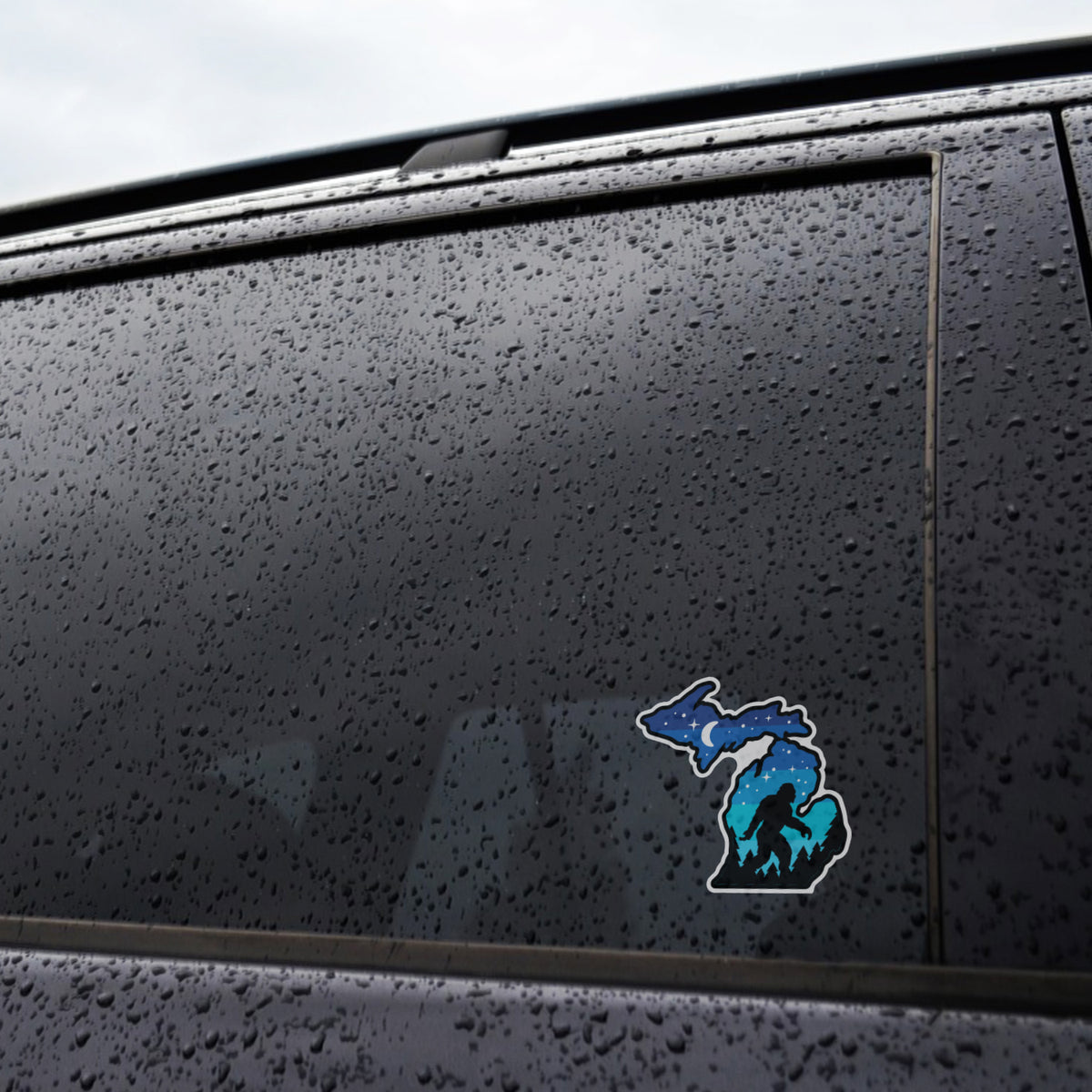 Raindrops cover a dark car window where the Michigan Sasquatch sticker sits near the lower corner of the glass.