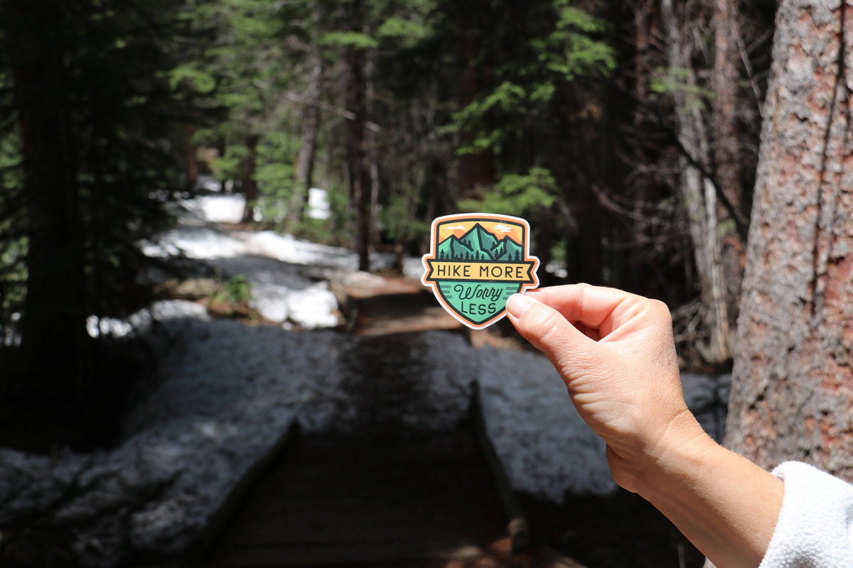 Beside a snowy creek and wooden steps, the Hike More Worry Less sticker is held out in one hand.
