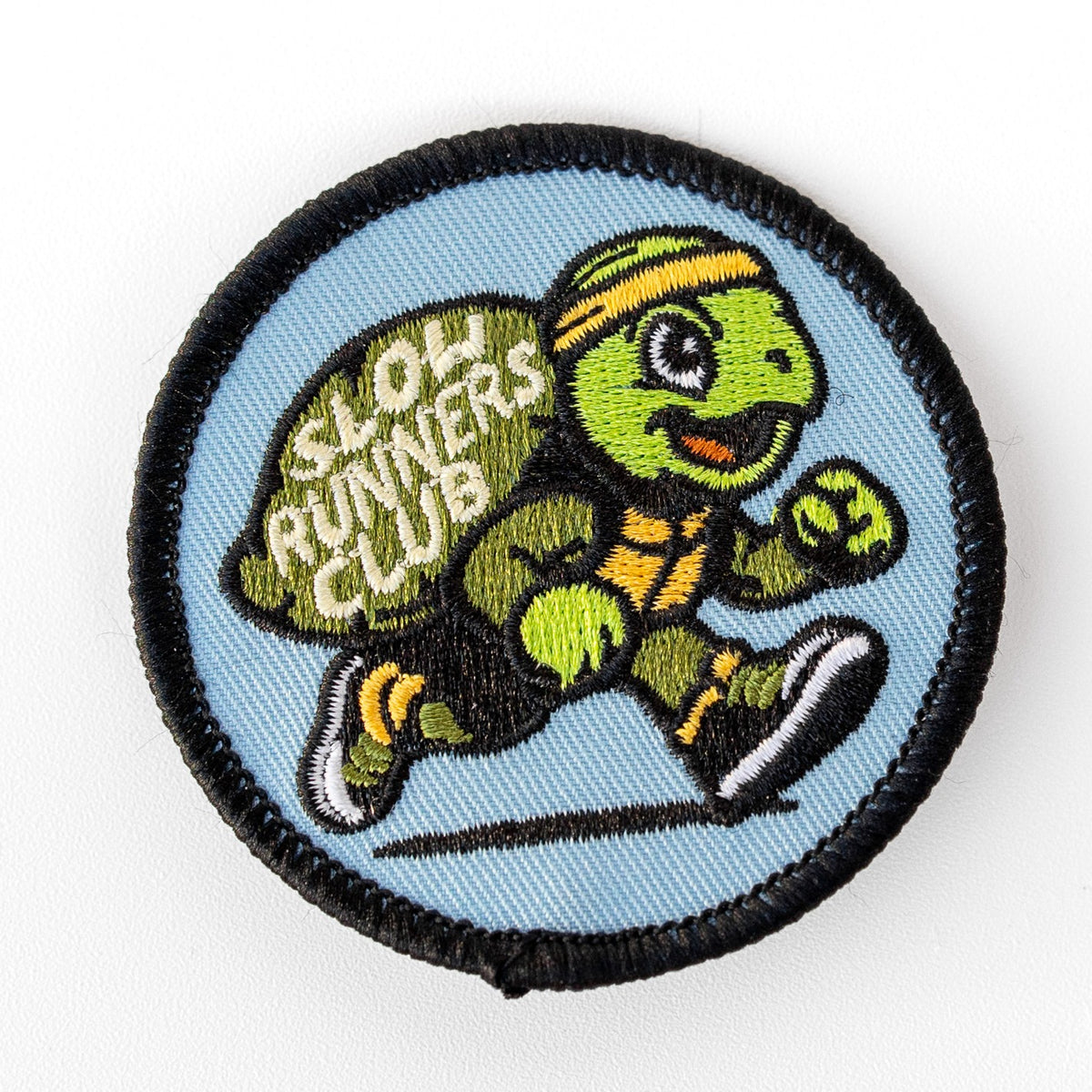 On a round blue background, the Slow Runners Club patch shows a jogging turtle in a yellow headband and sneakers.