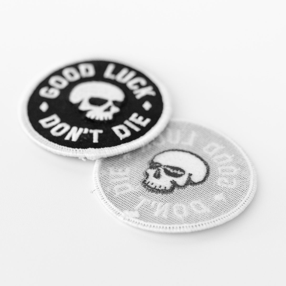 Two Good Luck Don't Die patches lean together, one showing the stitched skull front and one its pale gauzy backing.