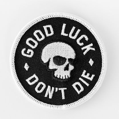 A round black patch shows a white skull with Good Luck Don't Die embroidered in white lettering around the edge.