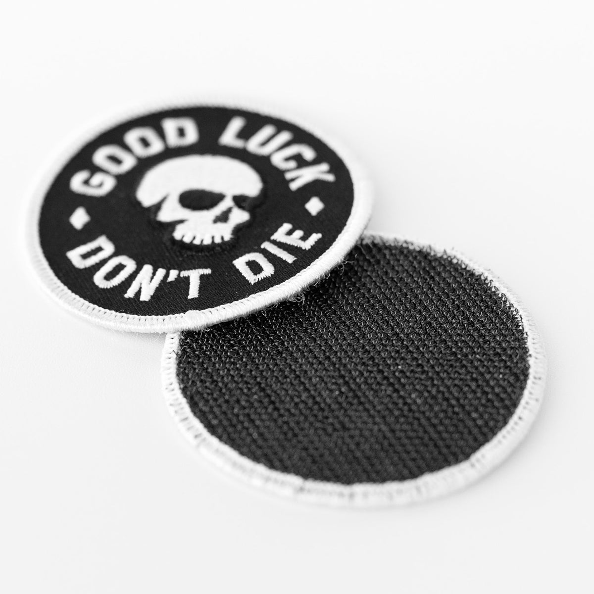One Good Luck Don't Die patch rests face up while a second behind it shows its plain black fabric backing.