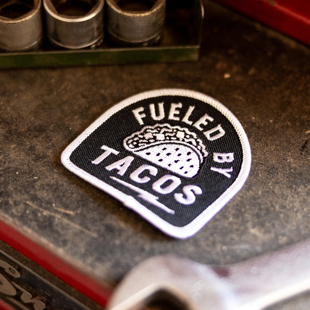 Resting on a grimy red toolbox beside sockets and a wrench, the Fueled By Tacos patch sits near a Snap-on logo.