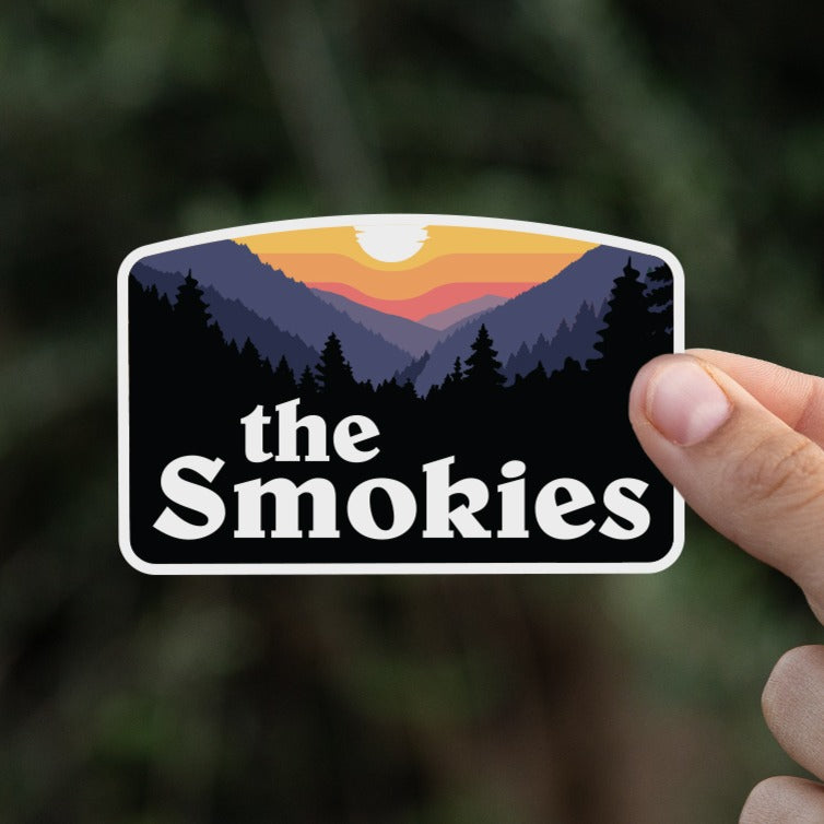 A hand holds the The Smokies sticker up against a blurred backdrop of dark green leaves.