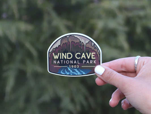A hand with a silver ring pinches the Wind Cave National Park sticker in front of a dense evergreen branch.