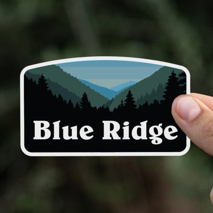 A hand holds the Blue Ridge sticker up against a blurred backdrop of dark green hedge branches.