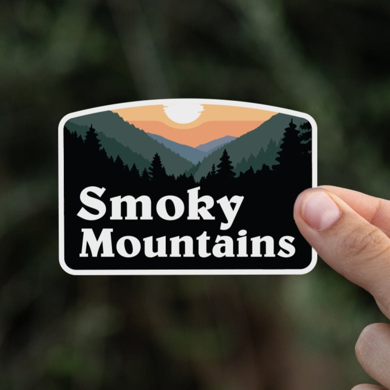 Pinched between two fingers, the small Smoky Mountains sticker stands out against dark blurred branches.