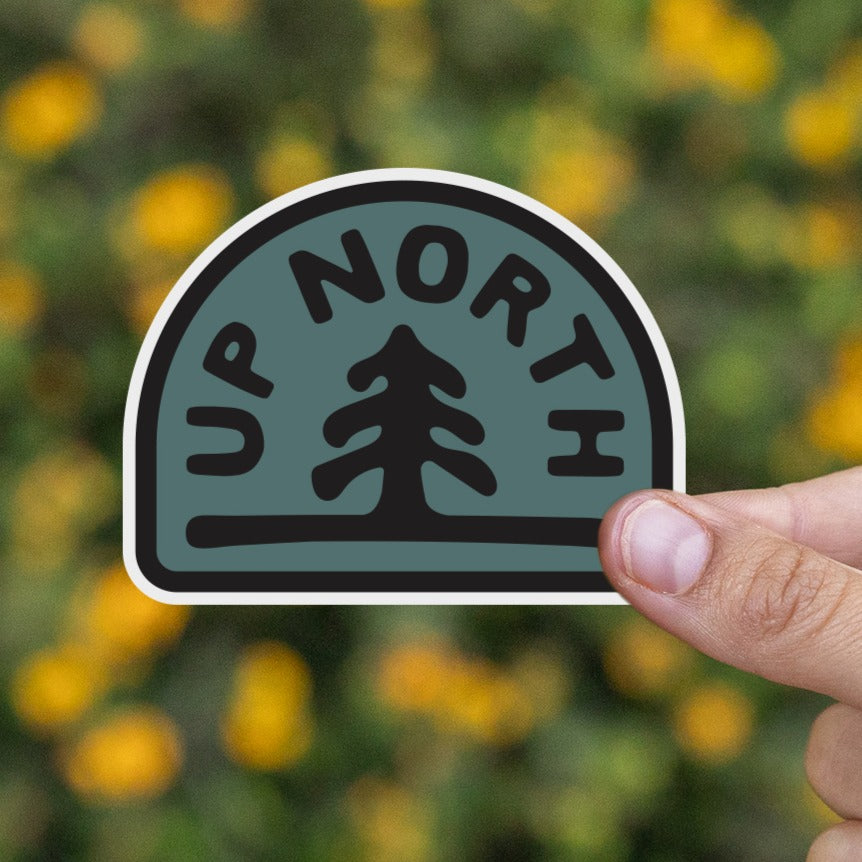 A hand presents the Up North sticker in front of a blurred field of small yellow wildflowers.