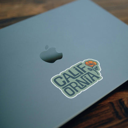 Stuck beside the logo on a grey laptop lid, the California Poppy sticker sits on a dark wood desk.