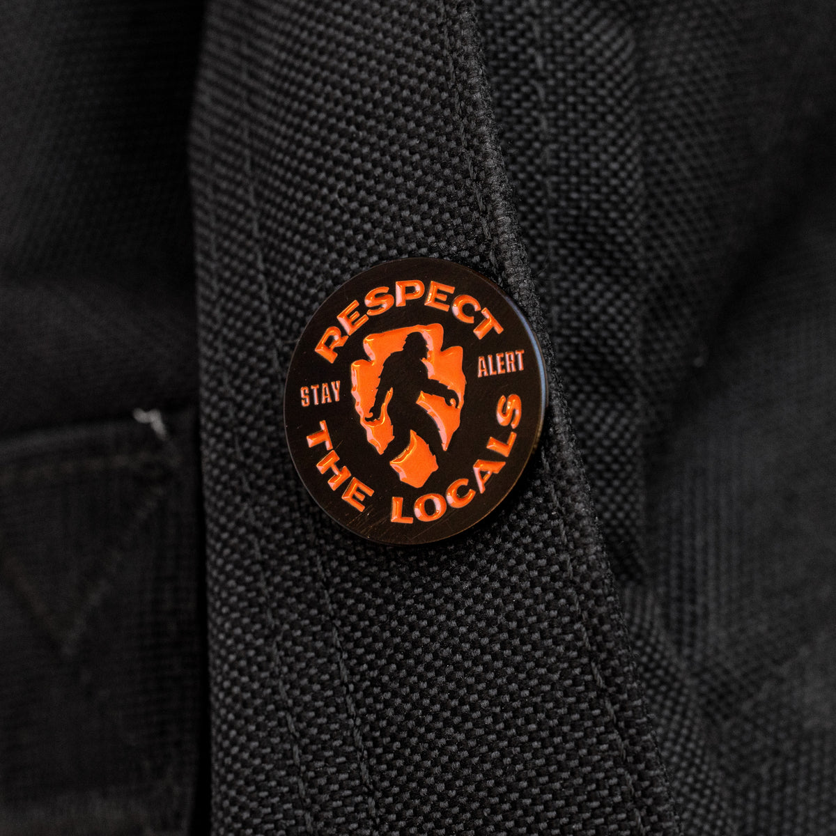 Pinned to a black backpack strap, the Respect The Locals pin shows its orange Bigfoot silhouette and Stay Alert text.