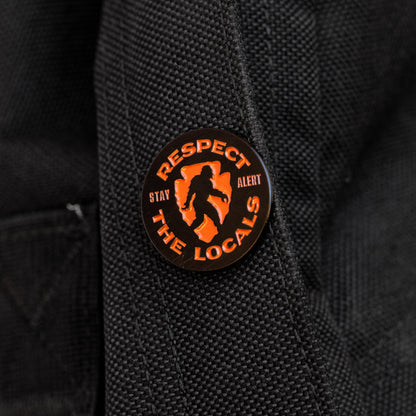 Pinned to a black backpack strap, the Respect The Locals pin shows its orange Bigfoot silhouette and Stay Alert text.
