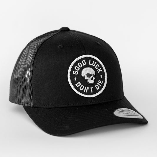 An all black Good Luck Don't Die trucker hat shows its skull patch clearly against the dark mesh and brim.