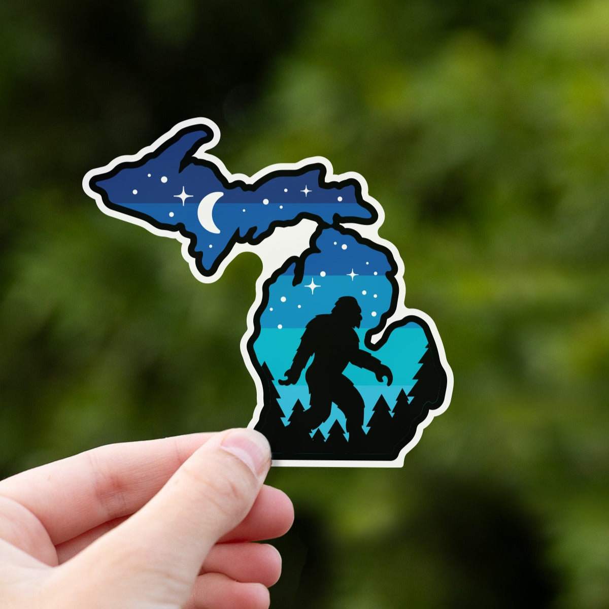 A hand holds the Michigan Sasquatch sticker up against a blurred backdrop of dark green leaves.