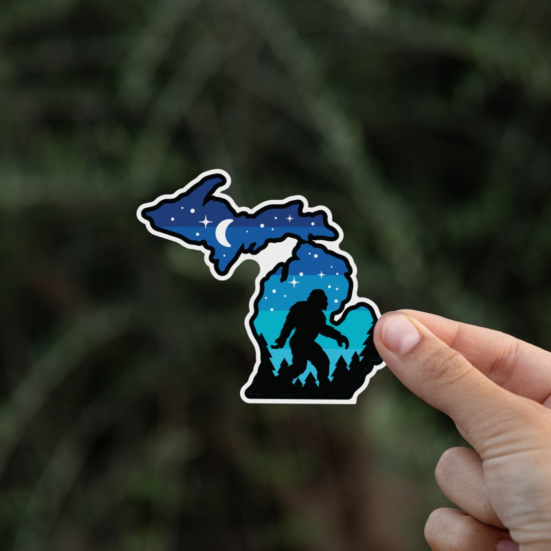 Pinched between two fingers, the Michigan Sasquatch sticker hovers in front of soft evergreen branches.