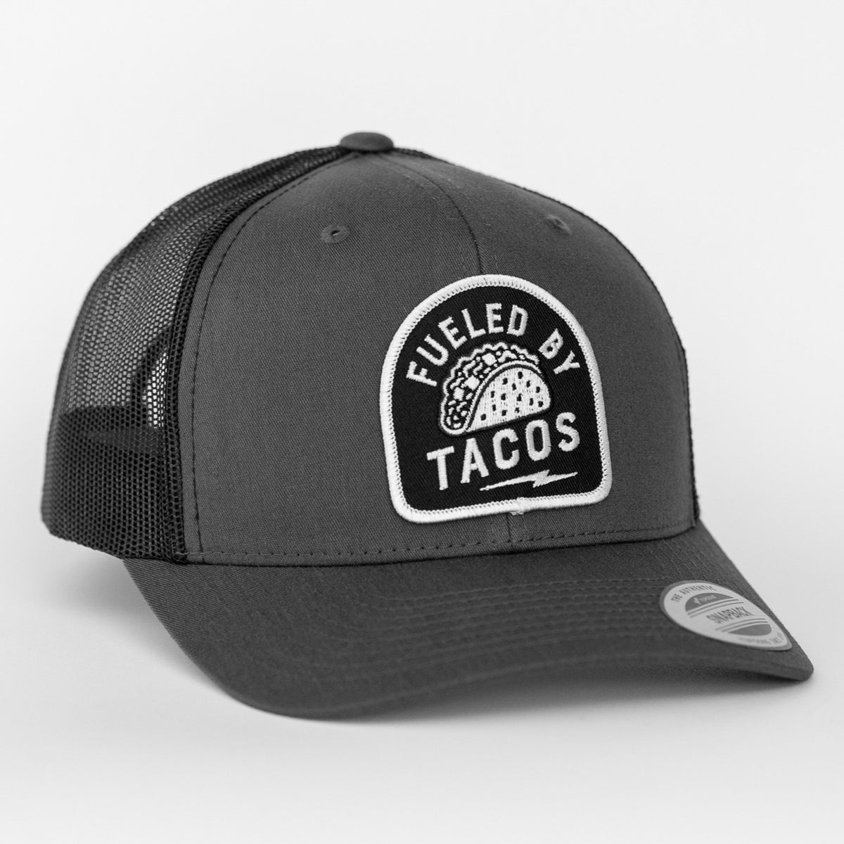 In charcoal grey with a black mesh back, the Fueled By Tacos trucker hat displays its taco patch on the curved brim side.