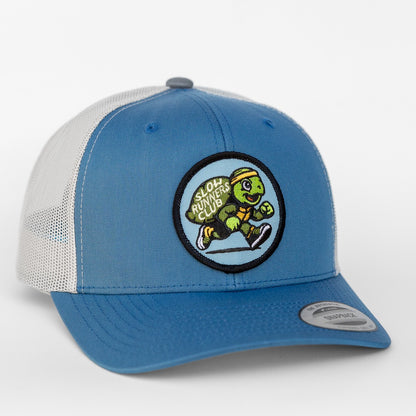 A blue and white Slow Runners Club trucker hat displays its round turtle patch angled toward the camera.